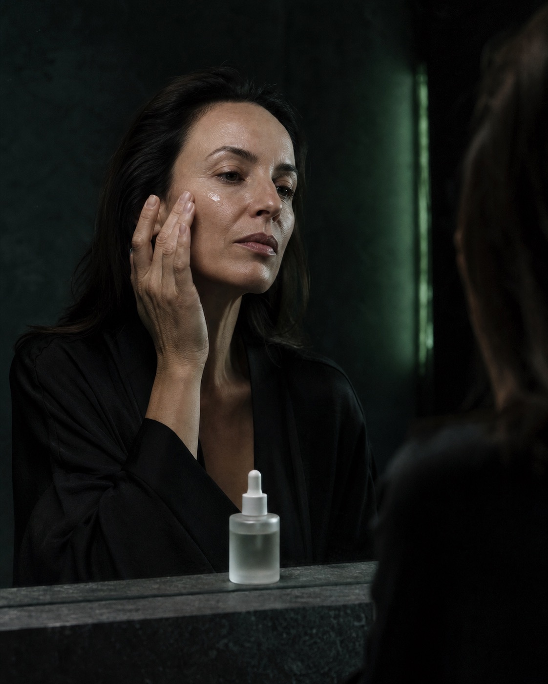 Woman applying clear facial serum at a dark bathroom mirror